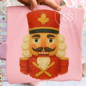 May include: A pink tote bag featuring a detailed illustration of a nutcracker soldier. The nutcracker is dressed in a red uniform with gold accents, a black mustache, and a white beard. The bag has the text "ORIGINAL" and "TSS THE SMILE SHOPPE CO."