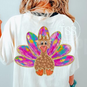 May include: Cream-colored t-shirt featuring a colorful turkey design. The turkey has a brown body with gold polka dots, a pink beak, and orange feet. The turkey's feathers are a mix of pink, blue, and yellow with gold accents. A pink cap is worn.