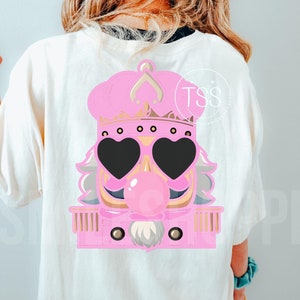 May include: Cream-coloured t-shirt with a pink cartoon nutcracker design. The design includes heart-shaped sunglasses, a bubblegum bubble, a crown, and the text "TSS".