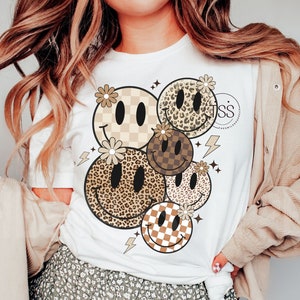 Op de afbeelding: Wit T-shirt met een grafisch ontwerp van vijf smiley-gezichten. Elk smiley-gezicht heeft een ander patroon, waaronder geruit, luipaardprint en bloemen.