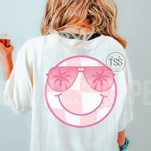 Peut inclure: T-shirt blanc avec un motif de visage souriant à carreaux rose et blanc. Le visage souriant porte des lunettes de soleil roses avec des palmiers reflétés dans les verres. Le texte "TSS" est visible dans le coin supérieur droit de l'image.