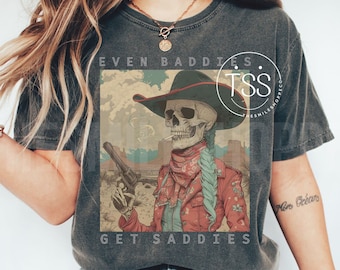 May include: A dark gray t-shirt with a vintage-style graphic of a skeleton wearing a cowboy hat and holding a gun. The text "Even Baddies Get Saddles" is printed below the graphic.
