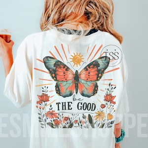 May include: A white t-shirt featuring a colourful butterfly graphic, a sun, and flowers. The text "be the good" is printed below the butterfly.