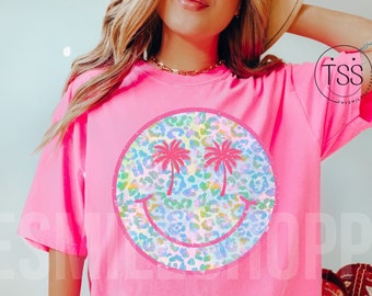 May include: Pink t-shirt with a large, colorful smiley face graphic. The smiley face is made of a leopard print pattern with palm trees for eyes.