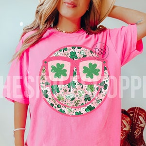 May include: Pink t-shirt with a green shamrock smiley face wearing sunglasses. The smiley face is made up of a pattern of shamrocks and hearts.