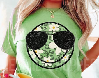 May include: A green t-shirt with a smiling face graphic. The face is surrounded by a leopard print and white daisies. The face is wearing sunglasses.