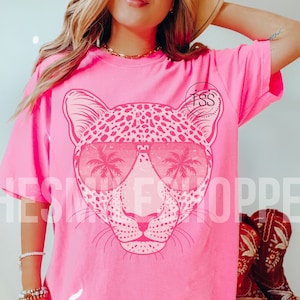 May include: Pink t-shirt with a graphic of a leopard wearing sunglasses with palm trees reflected in the lenses. The leopard is pink and white with a black outline.