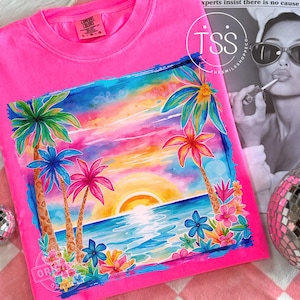 May include: A bright pink t-shirt featuring a colorful tropical sunset design. The graphic includes palm trees, ocean waves, and a vibrant sky with shades of orange, pink, and blue. The shirt has a crew neck and short sleeves.