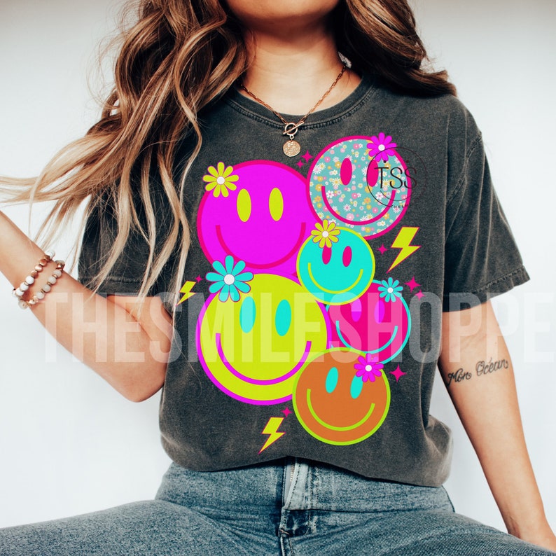 Op de afbeelding: Een donkergrijs T-shirt met een grafisch ontwerp van kleurrijke smiley-gezichten met bloemen en bliksemschichten. De tekst "The Smile Shop" is zichtbaar op de achtergrond.