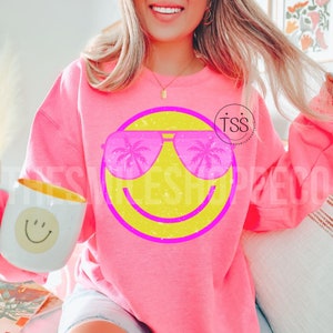 May include: A bright pink sweatshirt featuring a large yellow smiley face with pink sunglasses reflecting palm trees. The sweatshirt has a crew neck and long sleeves. A white mug with a smiley face is held in the left hand.