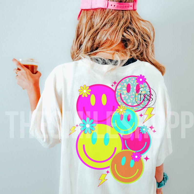 Op de afbeelding: Een wit T-shirt met een ontwerp van vijf kleurrijke smiley-gezichten met bloemen en bliksemschichten. De smiley-gezichten zijn in verschillende kleuren, waaronder roze, geel, blauw en oranje.