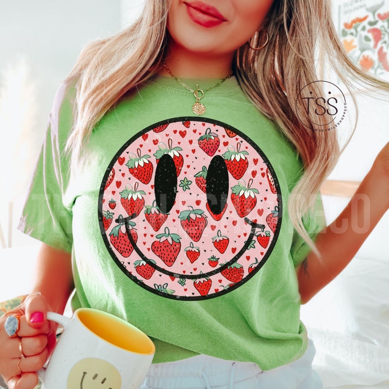 May include: Green t-shirt with a large smiley face graphic. The smiley face is filled with a pattern of pink hearts and red strawberries.