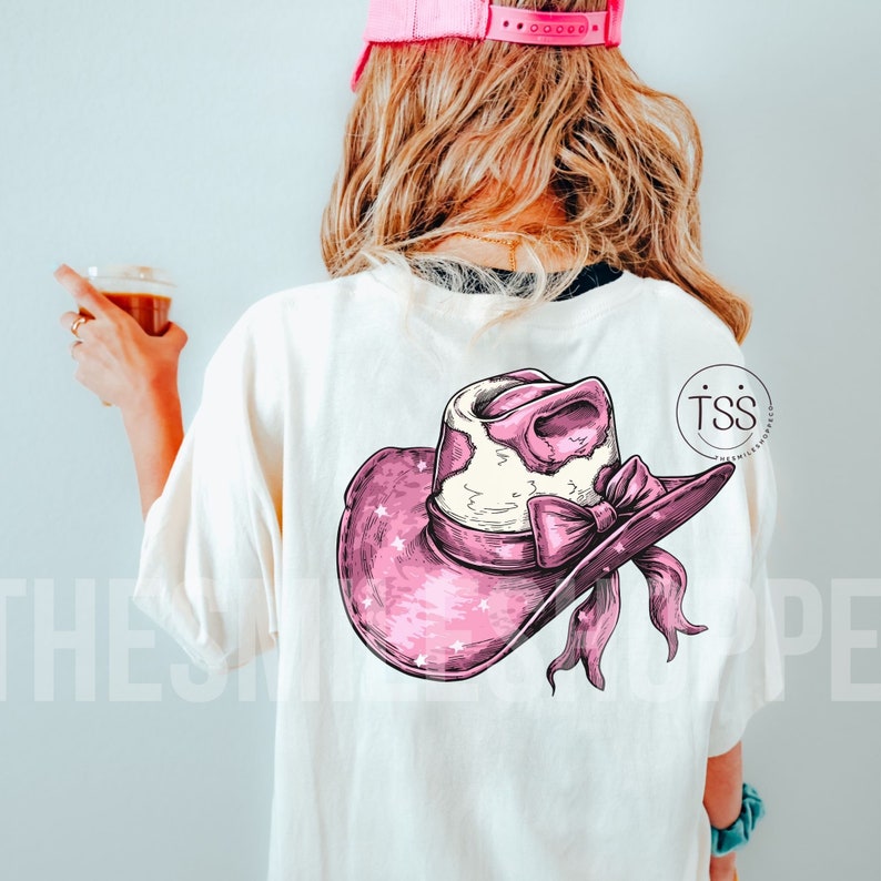 May include: A white t-shirt with a pink and white cowgirl hat graphic on the back. The hat has a white bow and pink stars.
