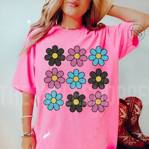 May include: Pink t-shirt with a graphic of nine black, pink, and blue glitter daisies.