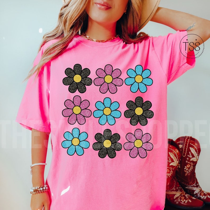 May include: Pink t-shirt with a graphic of nine black, pink, and blue glitter daisies.
