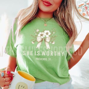 Puede incluir: Camiseta verde con un lazo blanco y un dise&ntilde;o floral. El texto "She is Worthy" y "Proverbs 31" est&aacute; impreso en la camiseta.