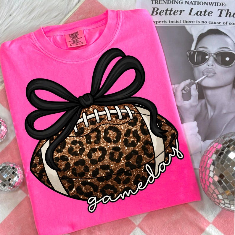 K&ouml;nnte beinhalten: Ein leuchtend pinkfarbenes Sweatshirt mit einem Football-Design, einer schwarzen Schleife und einem Leopardenmuster. Das Wort "gameday" ist in einer Schriftart unter dem Football geschrieben. Das Shirt ist von der Marke Comfort Colors.