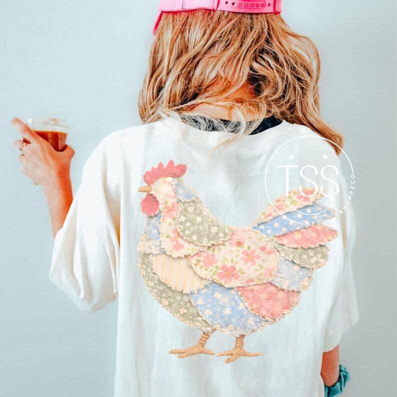May include: Cream-colored t-shirt featuring a patchwork chicken design on the back. The chicken is made of various pastel fabrics with floral patterns. The design includes a red comb and yellow feet. The shirt is worn by a person with blonde hair.