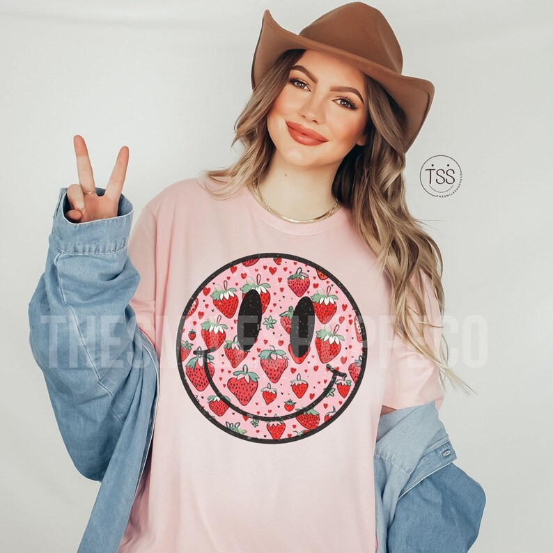 May include: A pink t-shirt with a large smiley face graphic. The smiley face is filled with a pattern of red strawberries and pink hearts.