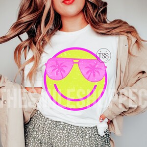 May include: White t-shirt featuring a neon yellow smiley face with pink sunglasses. The sunglasses have palm tree silhouettes in the lenses. The shirt also has the text "The Smile Shoppe" in a circle on the upper right.