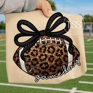 K&ouml;nnte beinhalten: Eine beigefarbene Tragetasche mit einem Football-Design mit Leopardenmuster und einer schwarzen Schleife. Das Wort "gameday" ist in Schreibschrift geschrieben. Der Hintergrund zeigt ein gr&uuml;nes Feld.