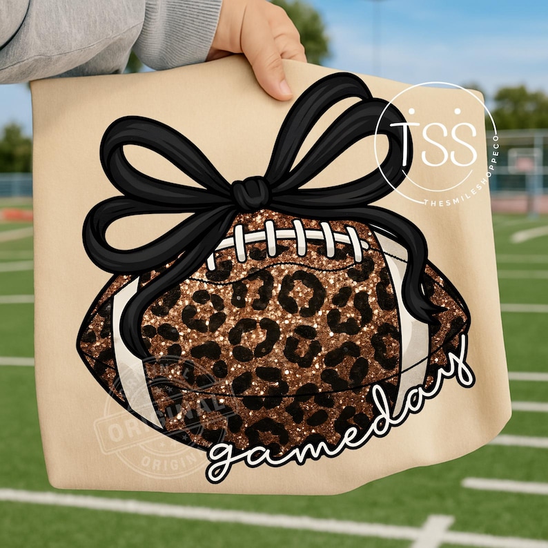 K&ouml;nnte beinhalten: Eine beigefarbene Tragetasche mit einem Football-Design mit Leopardenmuster und einer schwarzen Schleife. Das Wort "gameday" ist in Schreibschrift geschrieben. Der Hintergrund zeigt ein gr&uuml;nes Feld.