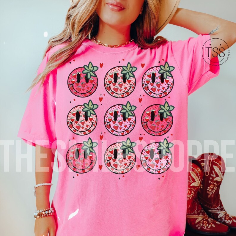 May include: Pink t-shirt with a pattern of smiling faces made of strawberries. Each face has a green leaf on top. The shirt has a short sleeve and a crew neck.