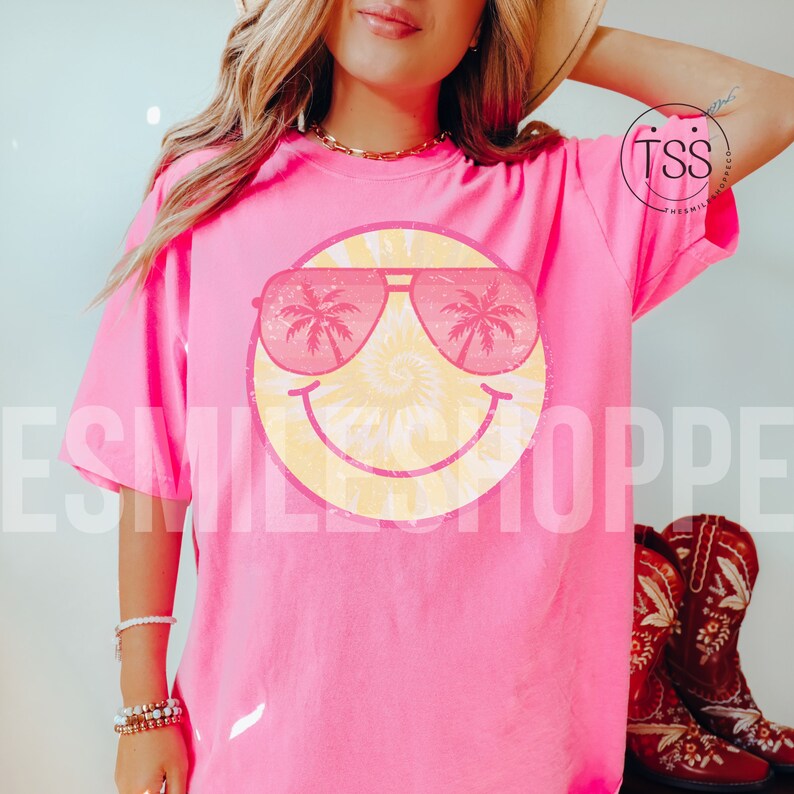 May include: Pink t-shirt with a yellow smiley face wearing pink sunglasses with palm trees on the lenses. The text "SMILESHOPPE" is visible in the background.