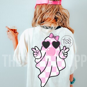 K&ouml;nnte beinhalten: Ein wei&szlig;es T-Shirt mit einem rosa-wei&szlig; karierten Geist, der herzf&ouml;rmige Sonnenbrillen und eine rosa Schleife tr&auml;gt. Der Geist macht mit beiden H&auml;nden Peace-Zeichen. Der Text "TSS" ist auf dem Shirt sichtbar.