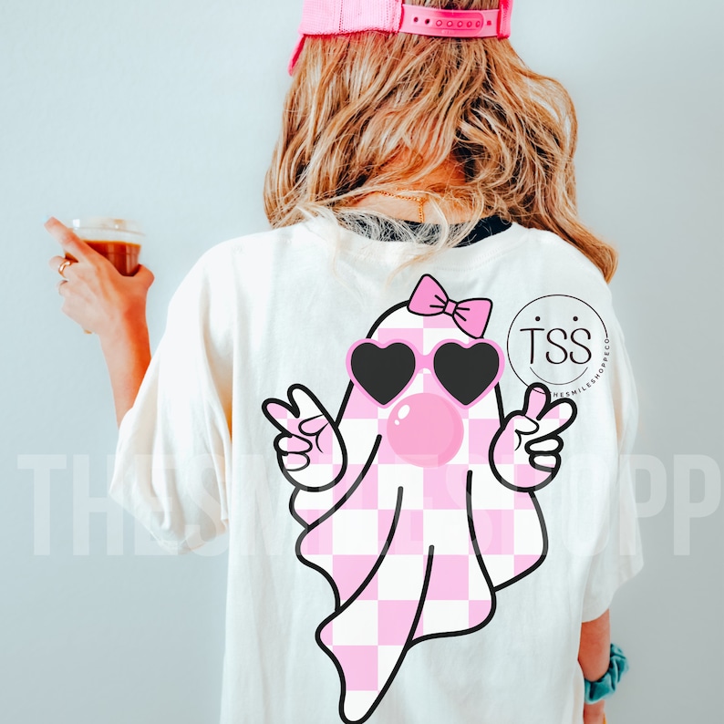 K&ouml;nnte beinhalten: Ein wei&szlig;es T-Shirt mit einem rosa-wei&szlig; karierten Geist, der herzf&ouml;rmige Sonnenbrillen und eine rosa Schleife tr&auml;gt. Der Geist macht mit beiden H&auml;nden Peace-Zeichen. Der Text "TSS" ist auf dem Shirt sichtbar.