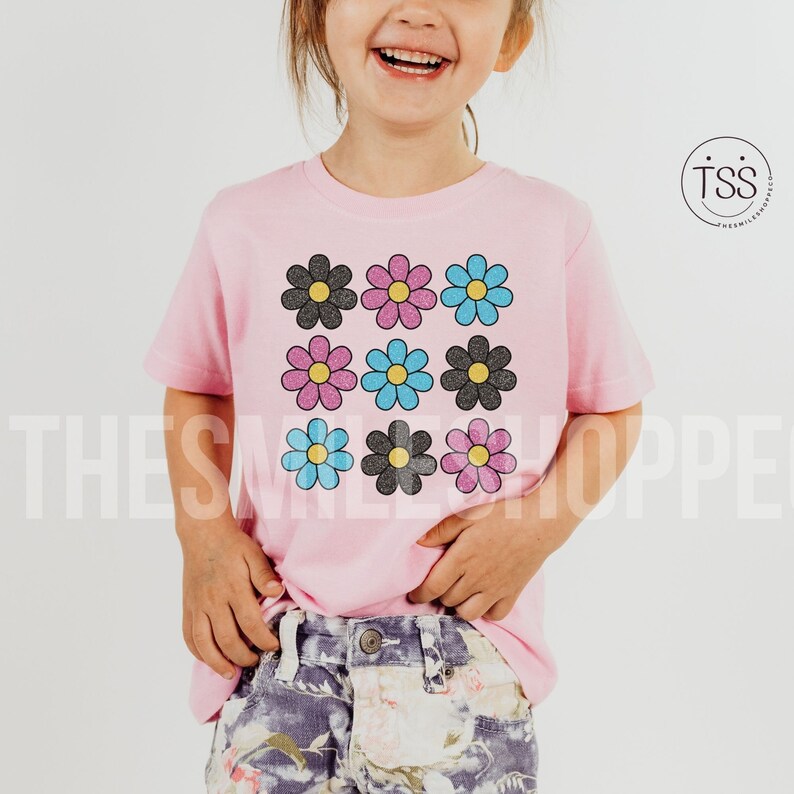 May include: A pink t-shirt with a graphic of nine colorful daisies. The daisies are arranged in a 3x3 grid pattern. The daisies are black, pink, blue, and light blue.