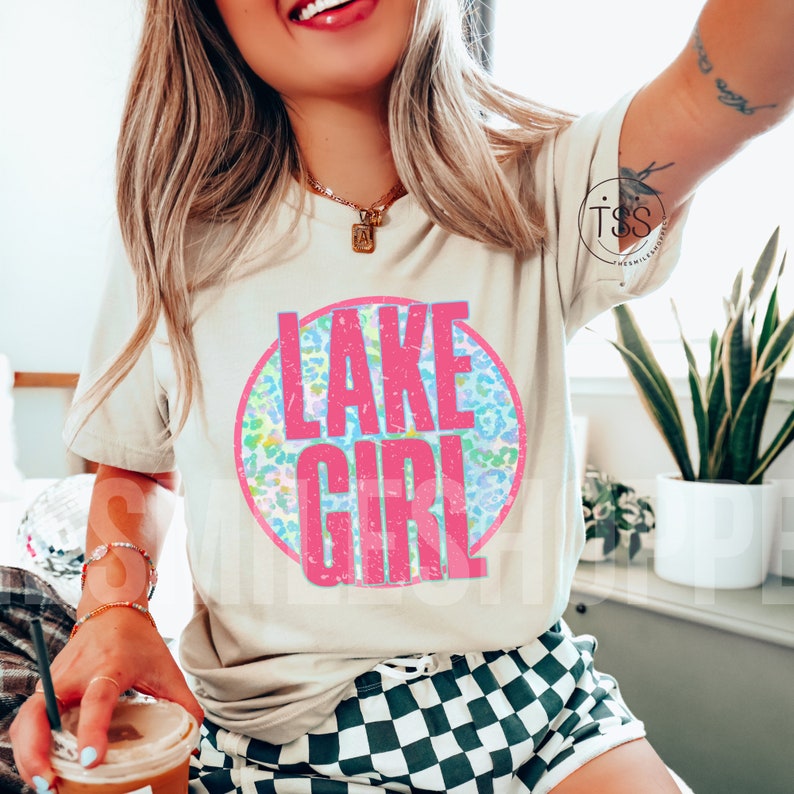 May include: A white t-shirt with a pink circle graphic that says "LAKE GIRL" in a distressed font. The circle graphic has a blue and green leopard print pattern.