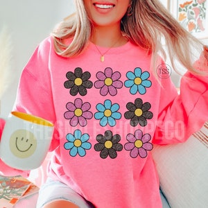 May include: Pink sweatshirt with a graphic of nine glittery daisy flowers in black, pink, and blue.