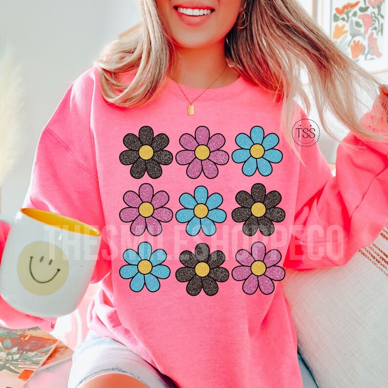 May include: Pink sweatshirt with a graphic of nine glittery daisy flowers in black, pink, and blue.