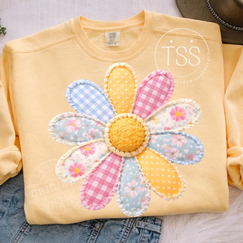 May include: A light yellow sweatshirt featuring a large, appliqued daisy. The daisy petals are a mix of patterns: blue gingham, pink gingham, floral, and yellow polka dots. The center of the flower is a textured yellow circle.