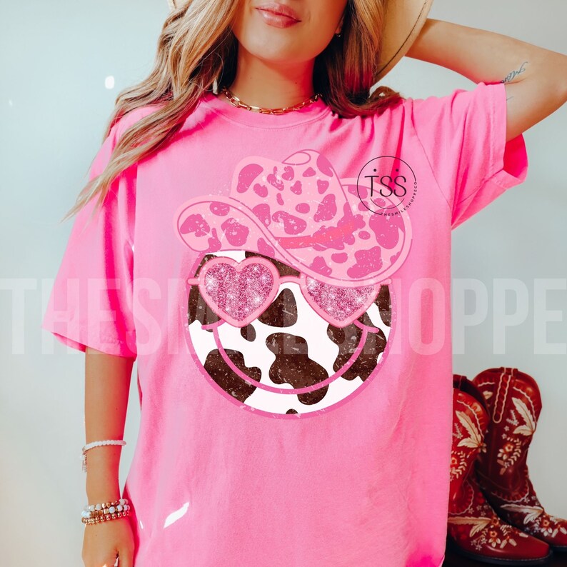 May include: Pink t-shirt with a cow print smiley face wearing a pink and white cow print cowboy hat and pink glitter heart sunglasses. The text "TSS" is visible on the shirt.