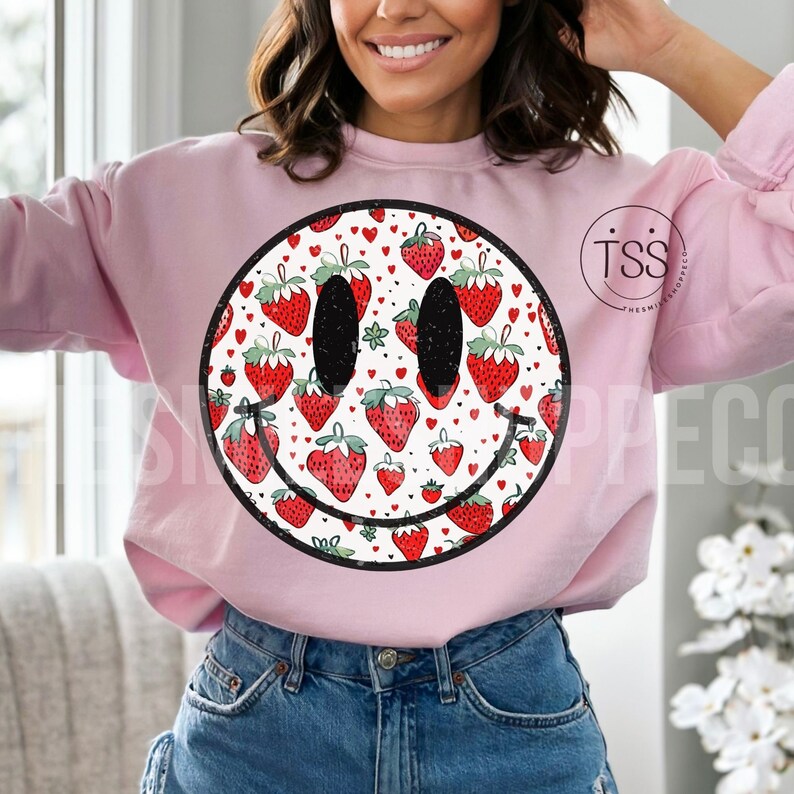 May include: Pink sweatshirt with a large smiley face graphic. The smiley face is filled with a pattern of red strawberries and white hearts.