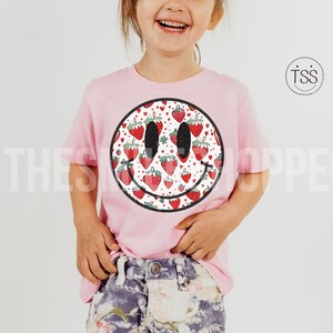 May include: A pink t-shirt with a white smiley face design. The smiley face is filled with a pattern of red strawberries.