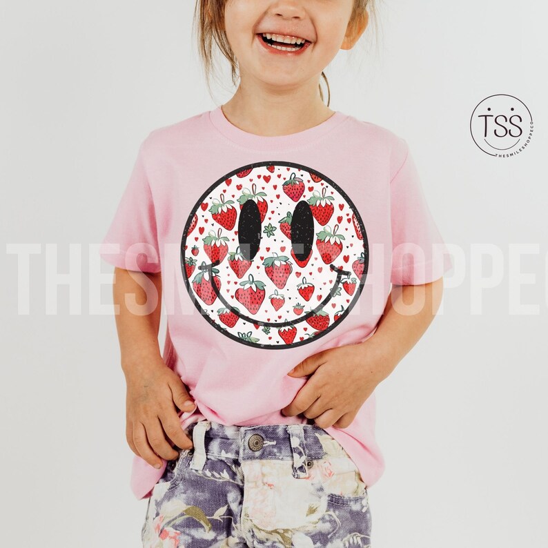 May include: A pink t-shirt with a white smiley face design. The smiley face is filled with a pattern of red strawberries.