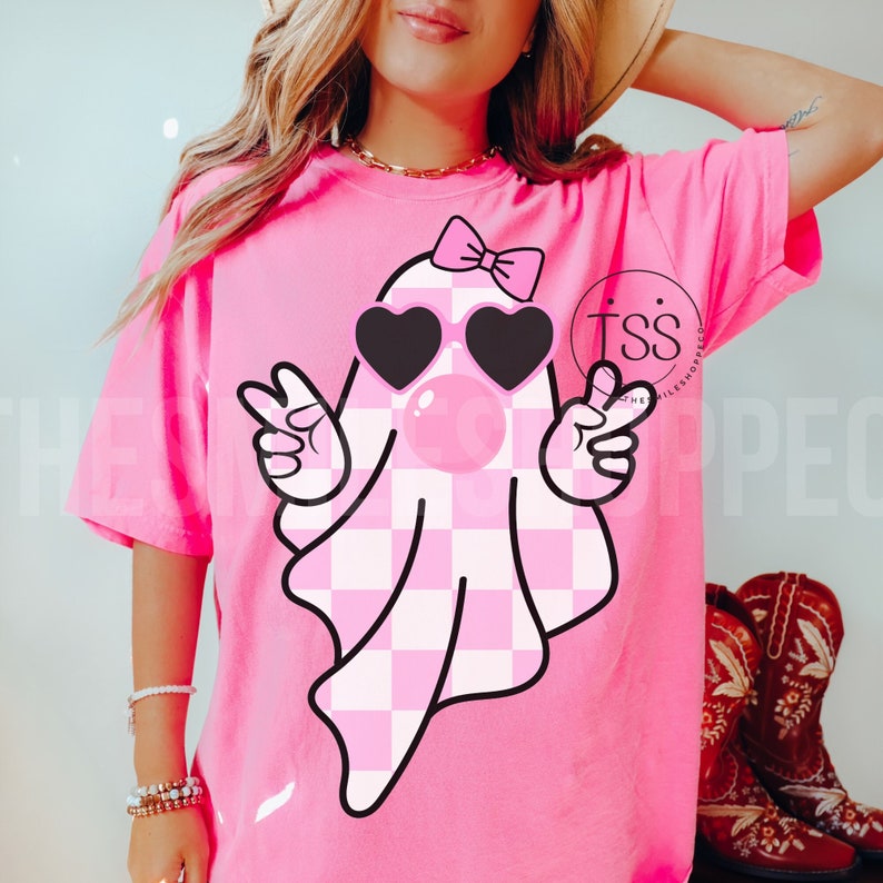 K&ouml;nnte beinhalten: Pinkes T-Shirt mit einem rosa-wei&szlig; karierten Geist, der eine Sonnenbrille und eine rosa Schleife tr&auml;gt. Der Geist bl&auml;st eine Blase.
