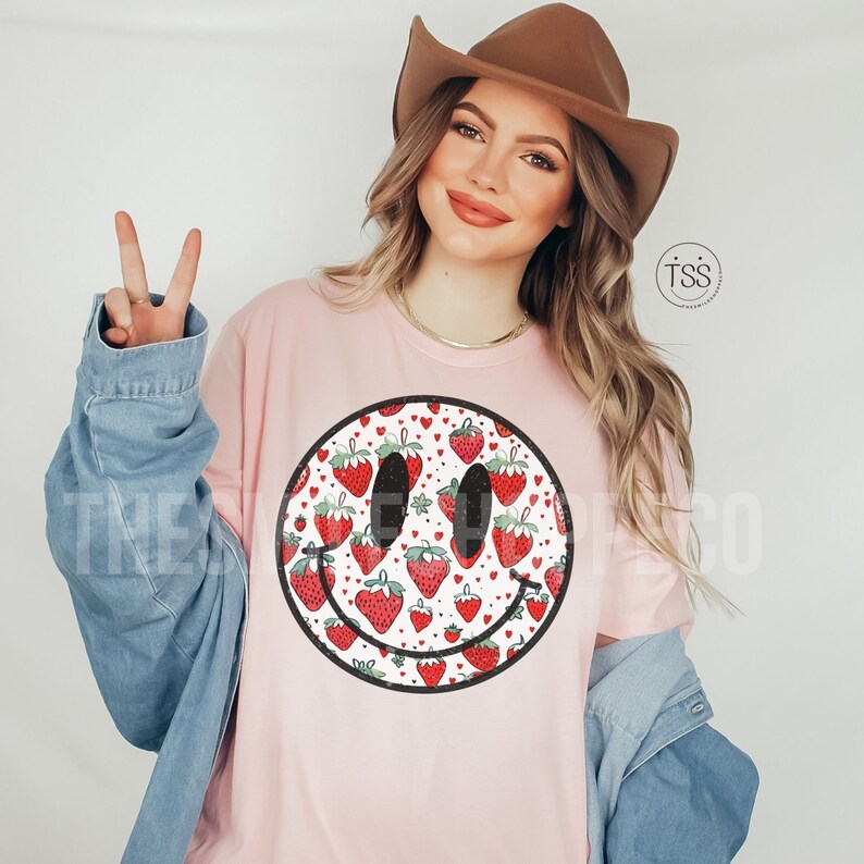 May include: A pink t-shirt with a smiley face design. The smiley face is filled with a pattern of red strawberries and white hearts.