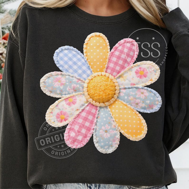 May include: Black sweatshirt featuring a large, colorful daisy applique. The daisy has yellow, pink, blue, and white petals with various patterns, including polka dots, gingham, and floral prints. The center of the flower is textured.