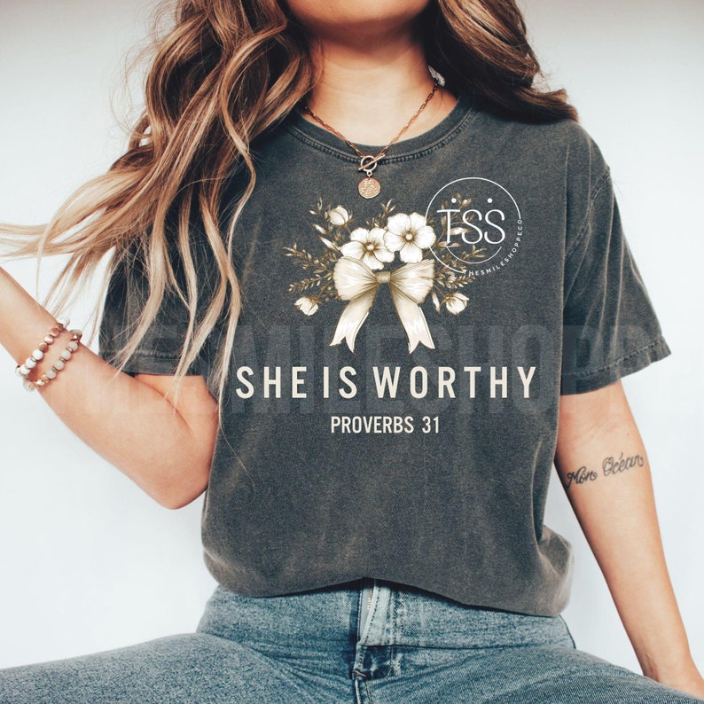 Puede incluir: Una camiseta gris con un dise&ntilde;o floral y un lazo. El texto "She is Worthy" y "Proverbs 31" est&aacute; impreso en la camiseta.