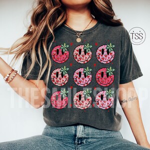 May include: A gray t-shirt with a pattern of nine smiley faces. Each smiley face is filled with a pattern of pink and red strawberries. The smiley faces have green leaves for eyes.