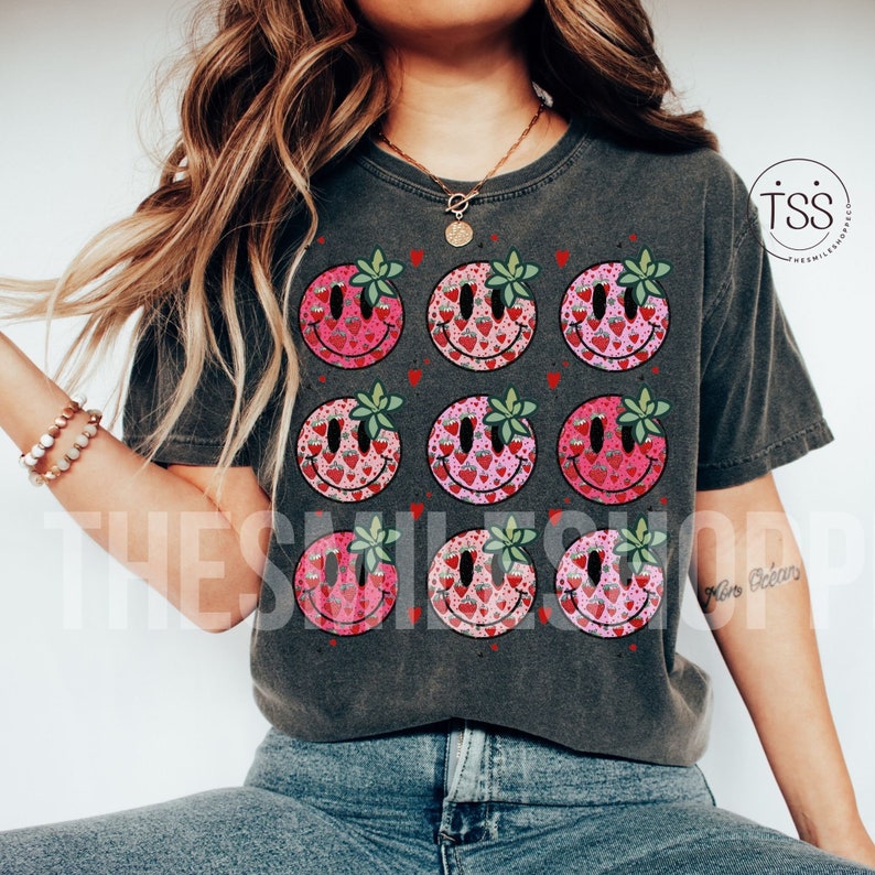 May include: A gray t-shirt with a pattern of nine smiley faces. Each smiley face is filled with a pattern of pink and red strawberries. The smiley faces have green leaves for eyes.