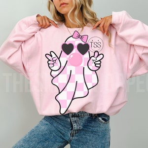 K&ouml;nnte beinhalten: Rosaer Pullover mit einem rosa-wei&szlig; karierten Geist, der eine Sonnenbrille und eine rosa Schleife tr&auml;gt. Der Geist bl&auml;st eine Blase und zeigt mit beiden H&auml;nden das Peace-Zeichen. Der Text "TSS" befindet sich auf dem Pullover.