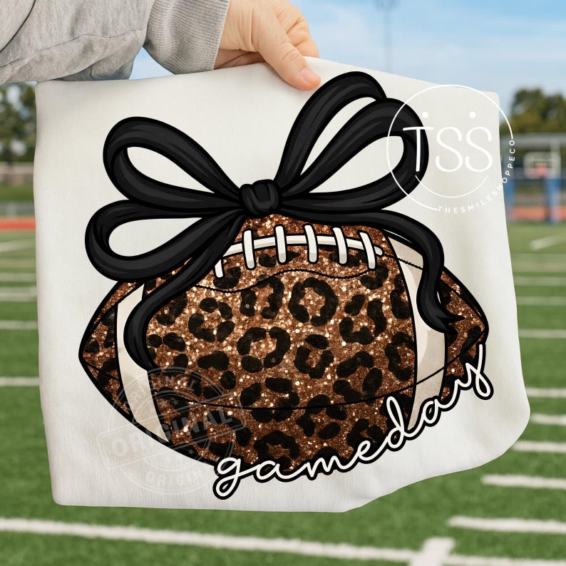 K&ouml;nnte beinhalten: Ein wei&szlig;es Sweatshirt mit einem Football-Design, das ein Leopardenmuster und eine schwarze Schleife aufweist. Die Worte "gameday" sind in Schreibschrift geschrieben. Das Design wird von einer Person gehalten.