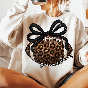 K&ouml;nnte beinhalten: Cremefarbenes Sweatshirt mit einem Football-Design mit Leopardenmuster und einer schwarzen Schleife. Das Wort "gameday" ist in einer Schreibschrift unter dem Football geschrieben. Ein l&auml;ssiges und sportliches Kleidungsst&uuml;ck.