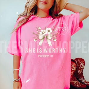Puede incluir: Camiseta rosa con un lazo blanco y un dise&ntilde;o floral. El texto "She is Worthy" y "Proverbs 31" est&aacute; impreso en la camiseta.