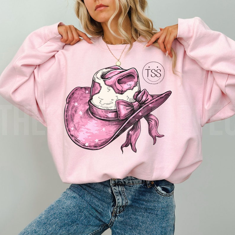 May include: Pink sweatshirt featuring a graphic of a pink cowboy hat with a white cowhide crown and a pink bow. The hat is decorated with white stars. The sweatshirt has the text "TSS" in a circle on the right side of the graphic.